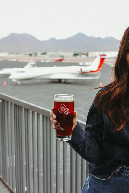 A guest on the Volanti patio holding a pint of amber beer, private jets and the McDowell Mountains behind