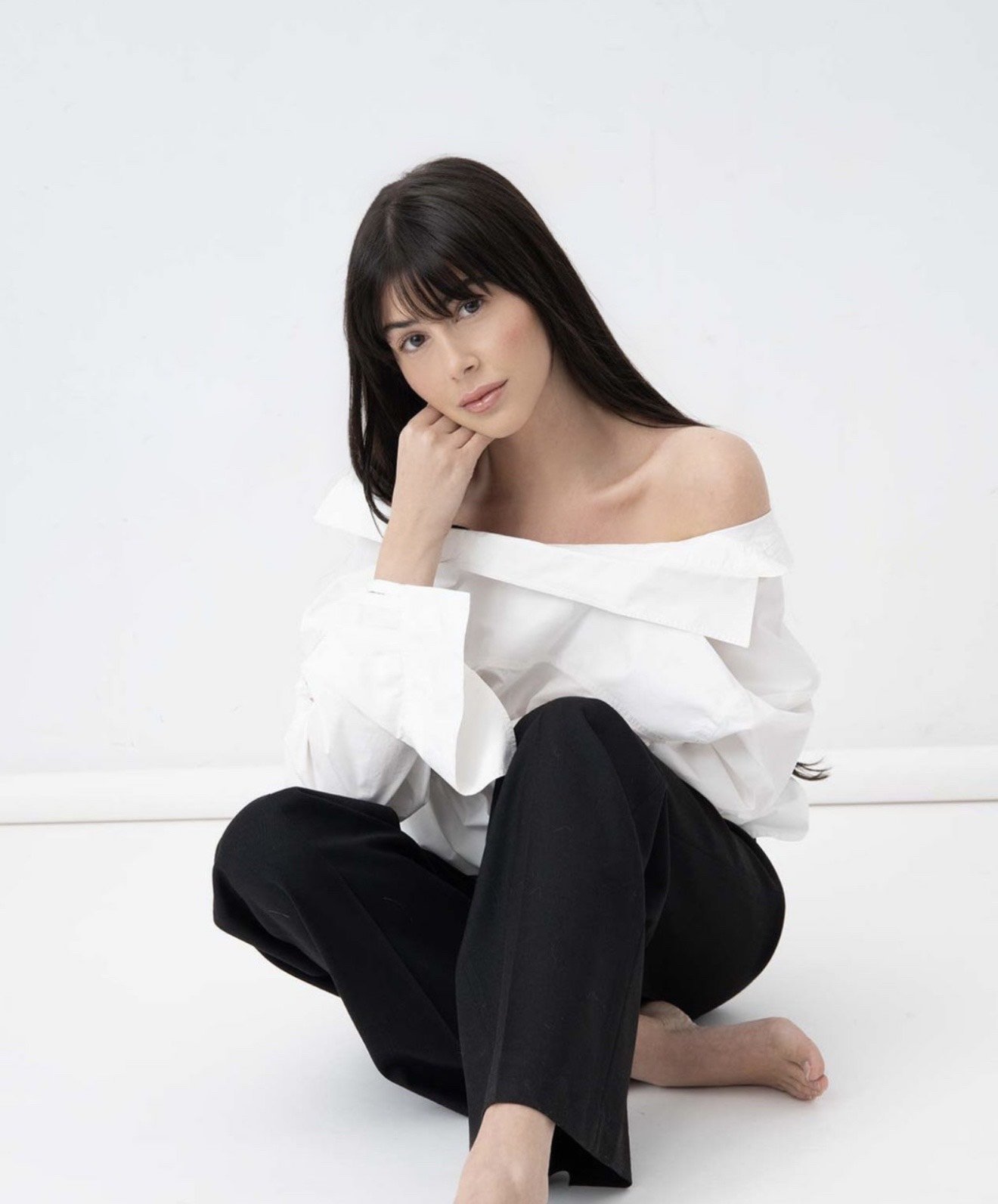 A woman sitting on the floor against a white background, wearing a white off-the-shoulder top and black pants, with dark hair and a neutral expression.