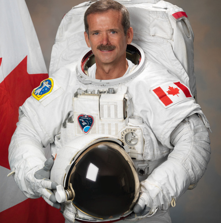 Astronaut in a white space suit holding a helmet, with a Canadian flag patch and a NASA badge, standing in front of a Canadian flag.