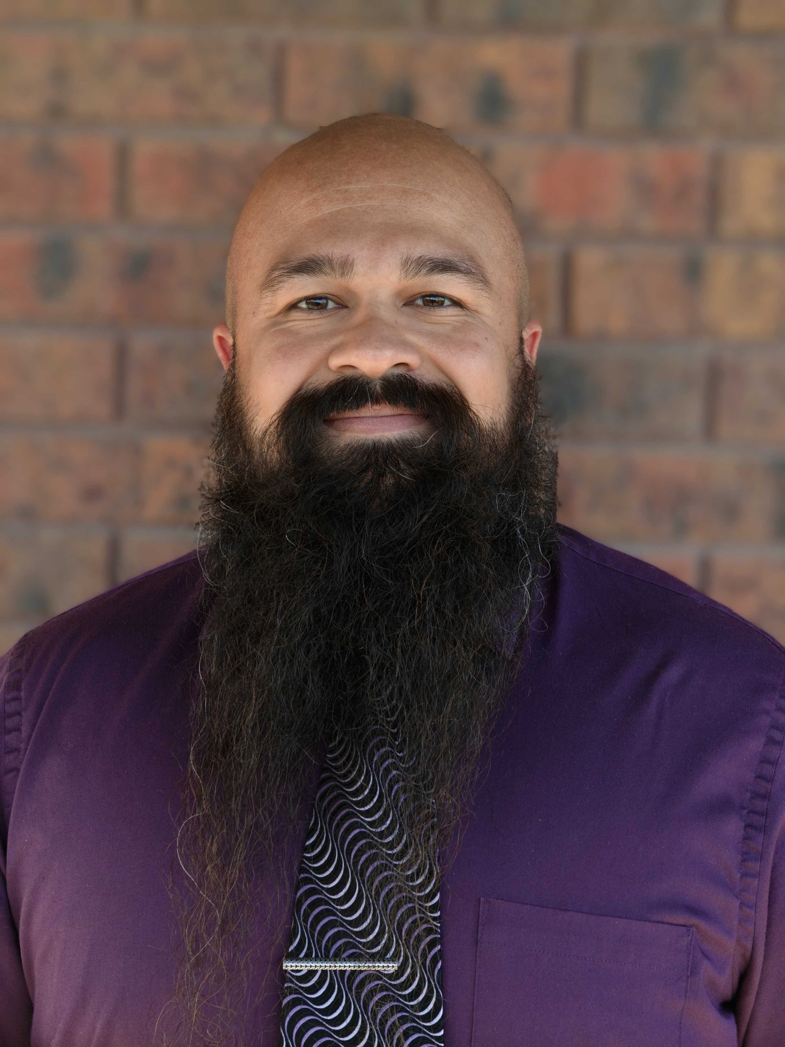 Portrait of a man with a full beard and a shaved head, wearing a purple shirt and a patterned tie, standing in front of a brick wall.