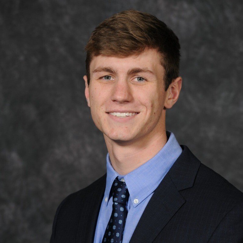 A young man with light brown hair, blue eyes, wearing a dark suit, light blue shirt, and patterned tie, smiling against a dark textured background.