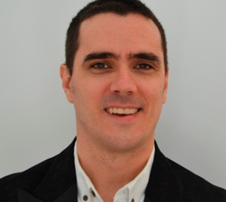 A young man with short dark hair, wearing a black blazer and a white shirt, smiling against a plain light background.
