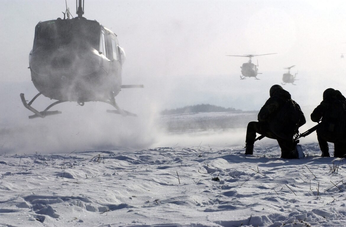 Two soldiers in winter gear kneeling on snow while three helicopters fly overhead in a snowy landscape.