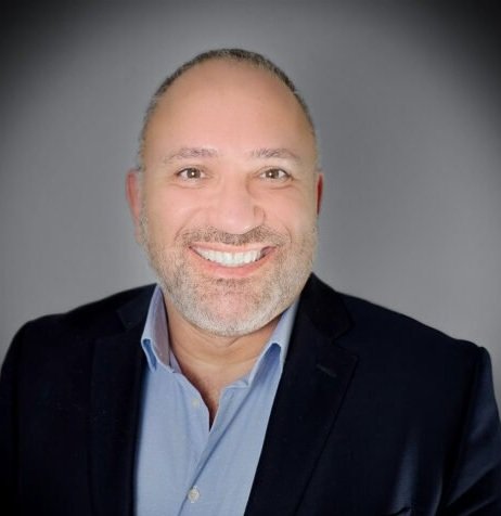 Headshot of a smiling man with short hair and a beard, wearing a blue dress shirt and a dark blazer, against a gray background.