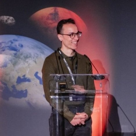 Person standing at a podium, smiling, with a backdrop of Earth and Mars images.