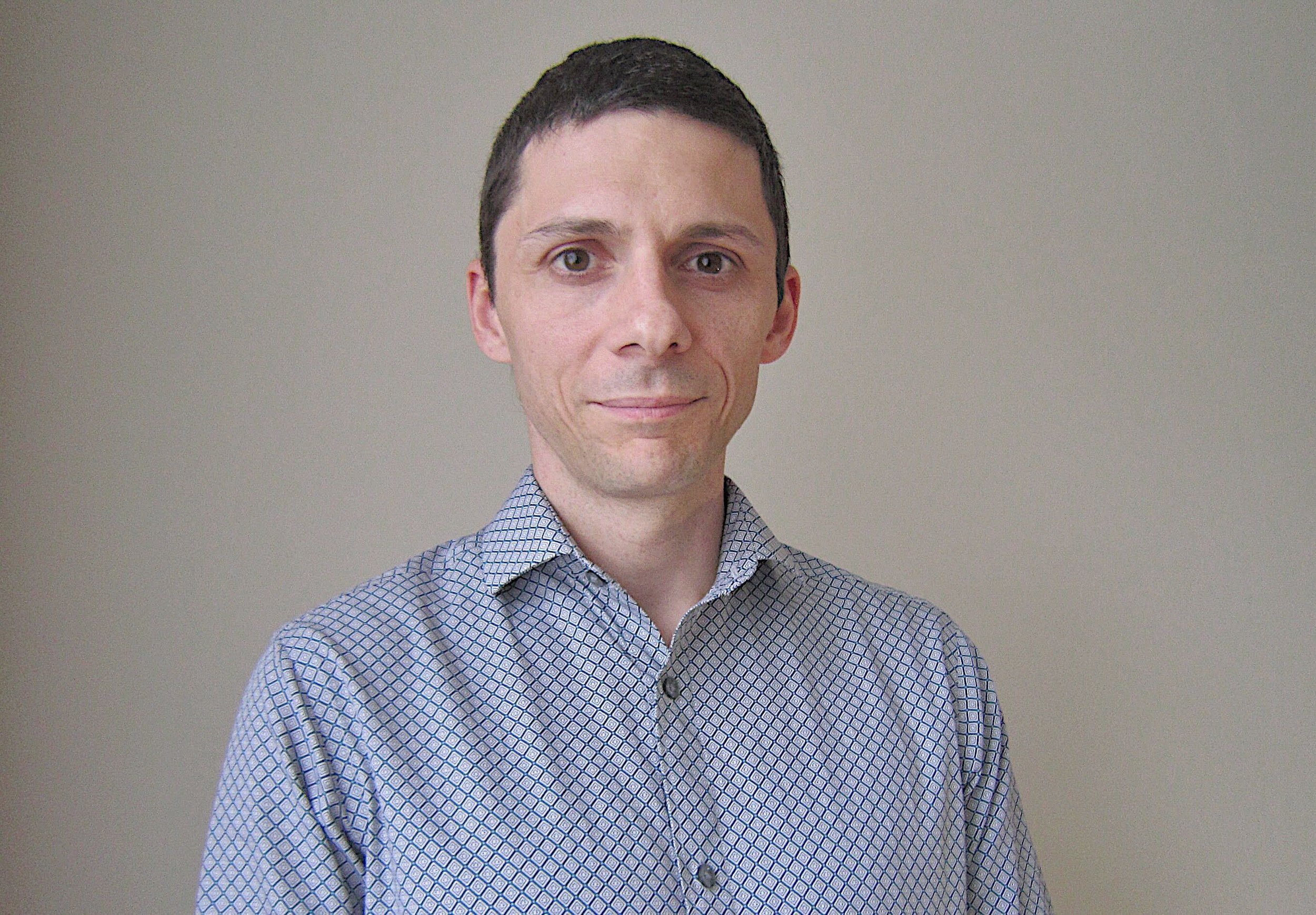 Portrait of a man with short dark hair, wearing a button-up shirt with a small diamond pattern, standing against a plain light-colored background.