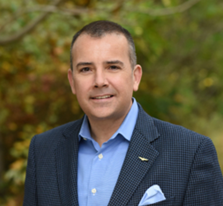 Man in blue suit and light blue shirt outdoors with green foliage background.