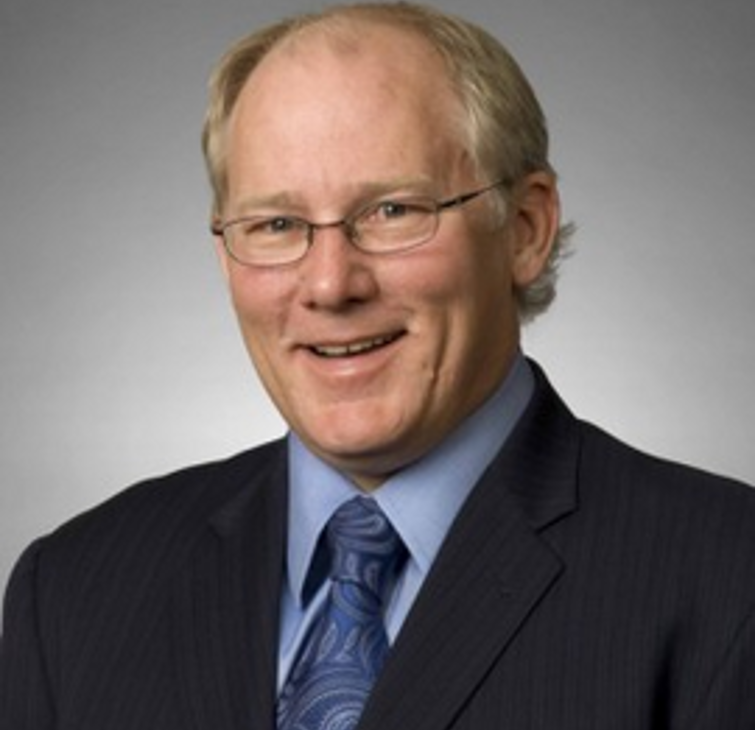 A middle-aged man with blond hair and glasses, wearing a dark pinstripe suit, light blue dress shirt, patterned blue tie, smiling against a neutral background.