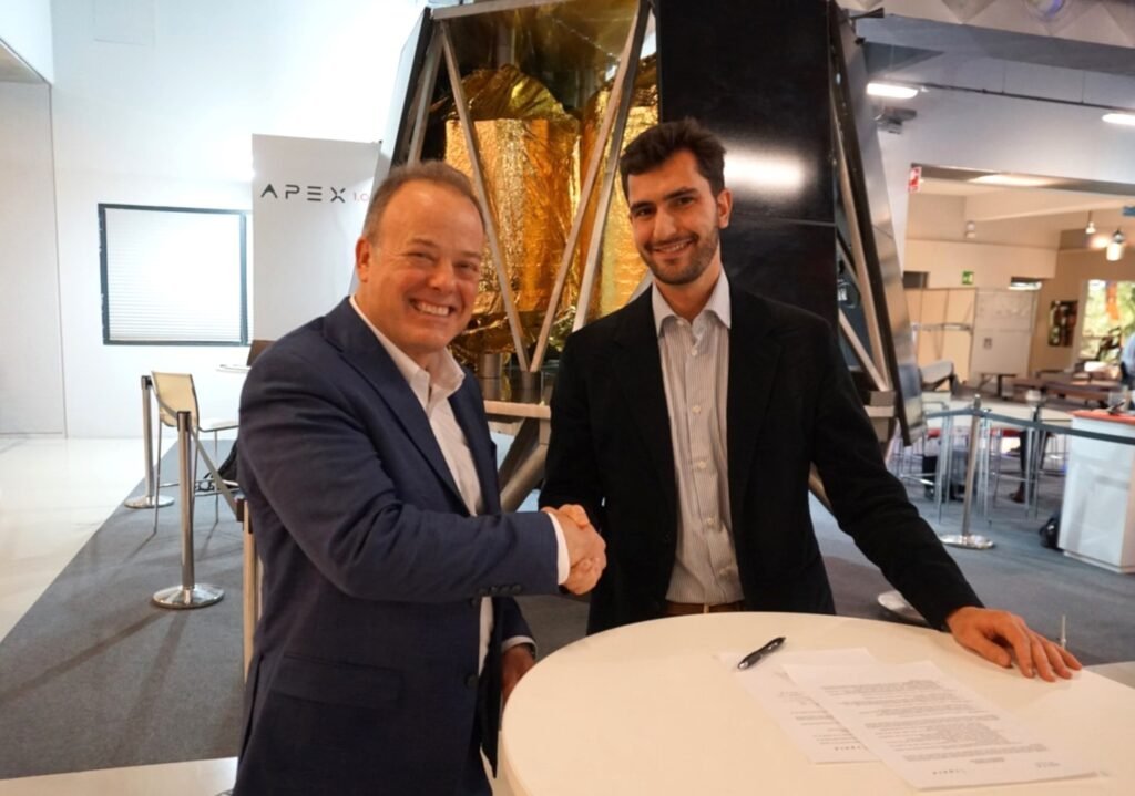 Two men in business suits shaking hands and smiling in a modern office or conference area, with a large gold and black mechanical structure in the background.
