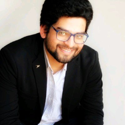 A man with dark hair, glasses, and a beard smiling, wearing a black blazer and a white shirt, against a neutral background.