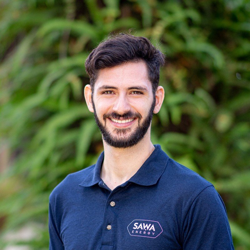 A young man with dark hair and a beard, smiling, wearing a navy blue polo shirt with a 'SAWA ENERGY' logo, standing outdoors with green foliage in the background.