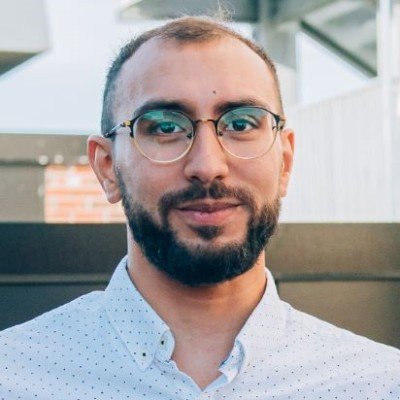 A man with glasses and a beard smiling outdoors, wearing a white collared shirt with small blue polka dots.
